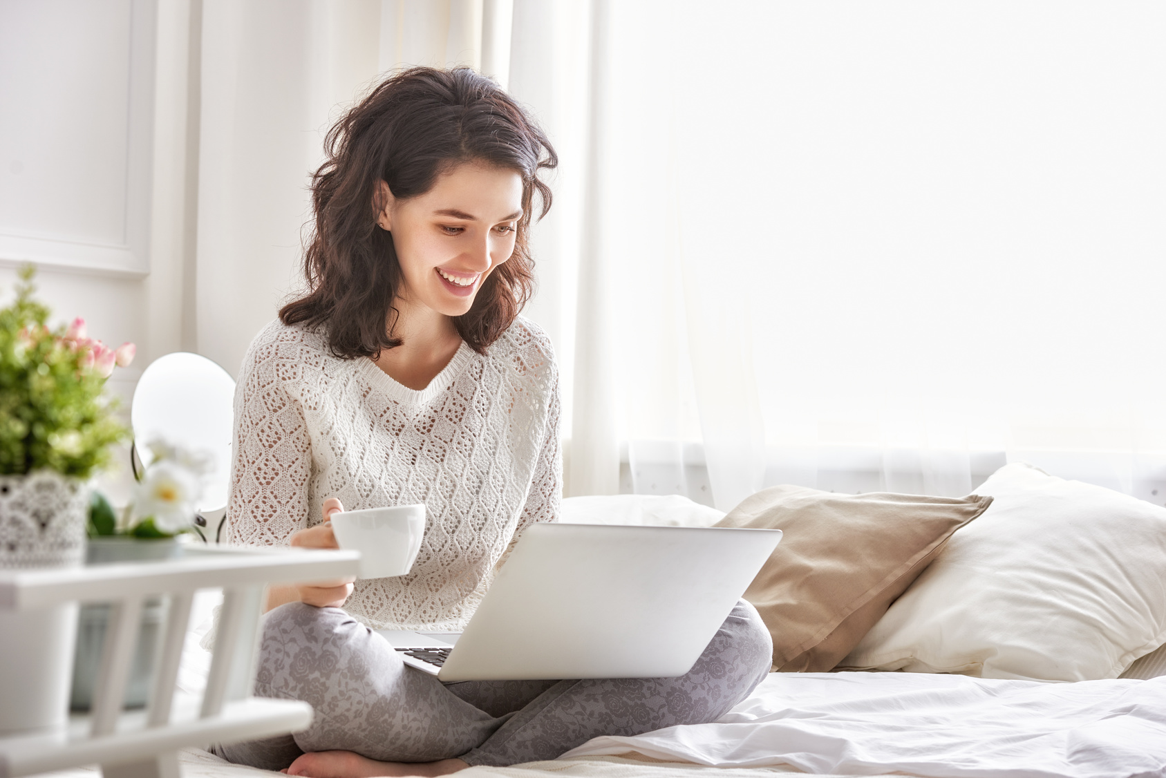 Eine Frau sitzt mit einem Laptop und einer Tasse auf einem Bett